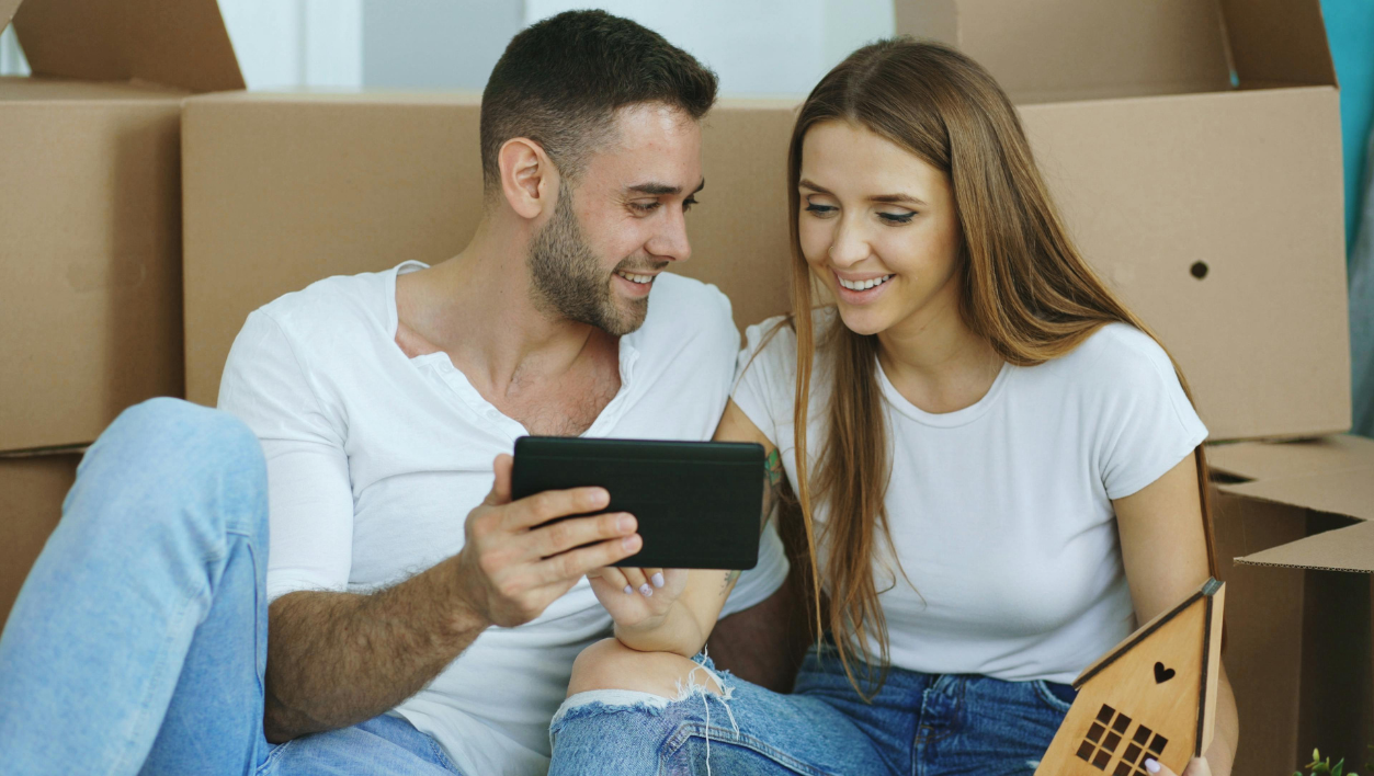 A couple sitting among moving boxes, looking at a tablet together